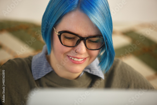 Nerdy white woman with blue hair working on laptop computer. Happy young adult person doing freelance work on notebook pc at home on lockdown. Cheerful female typing text with toothy smile