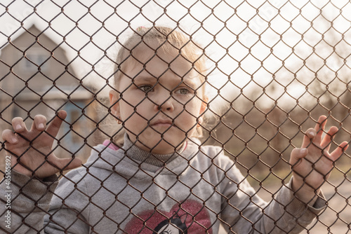A little girl with a sad look behind a metal fence. Social problem of refugees and forced migrants