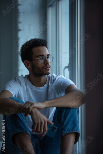 Attractive african american man in glasses and a white t-shirt sits on a windowsill.