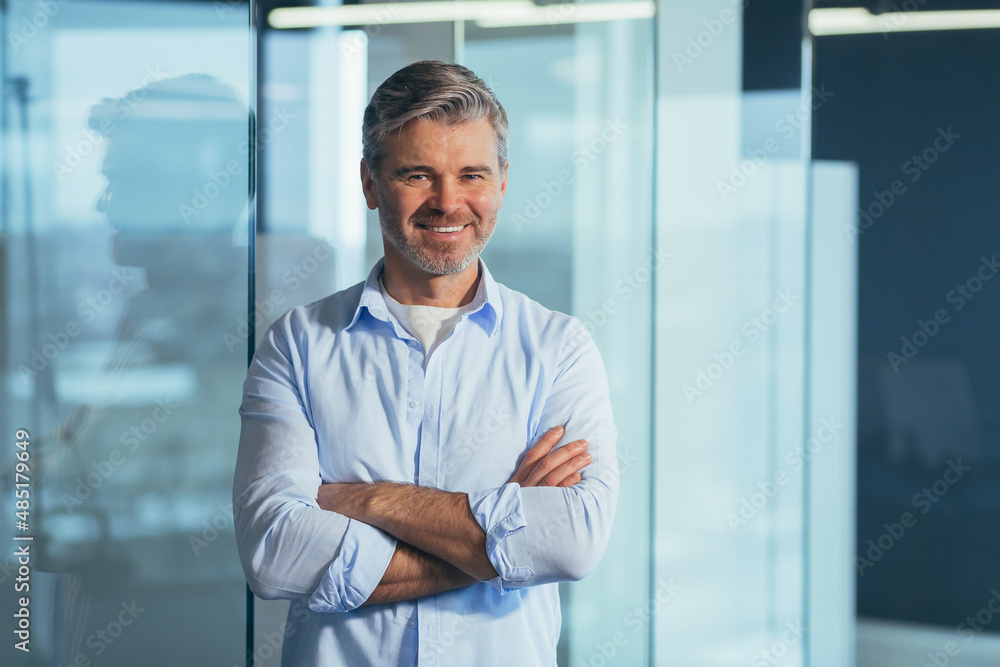 Portrait of a successful and happy gray-haired boss, a businessman in a modern office marveling at the camera and smiling with crossed arms Portrait of a successful and happy gray-haired boss, a businessman in a modern office marveling at the camera and smiling with crossed arms