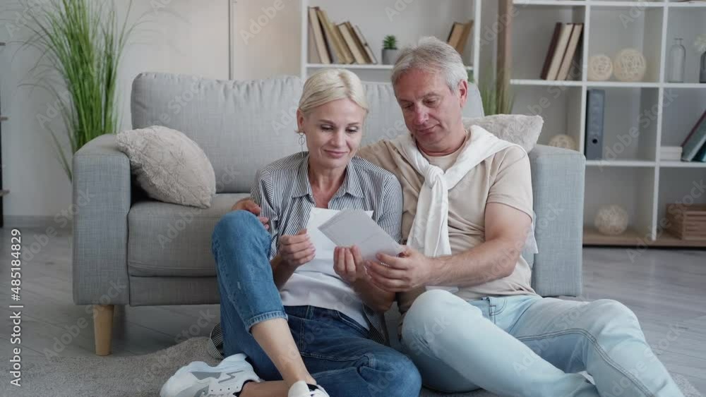 Photo memories. Happy couple. Leisure day. Casual middle-aged man and woman looking family pictures sitting floor together in light home interior.