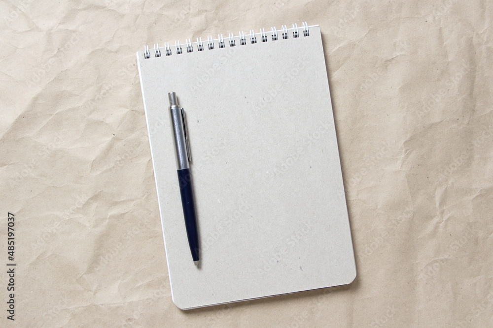Gray notepad with white coiled spring and pen on a background of beige crumpled craft paper. With empty space for text and design Gray notepad with white coiled spring and pen on a background of beige crumpled craft paper. With empty space for text and design