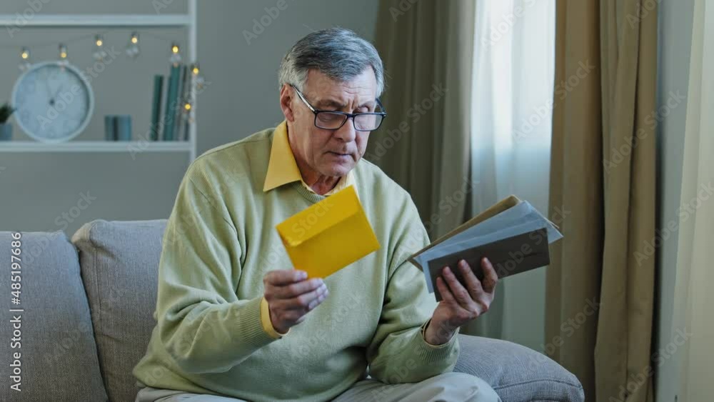 60s elderly man recipient with glasses sorting letters in living room concentrated mature grandfather sitting on sofa looking at correspondence notice bank account caucasian aged pensioner check bills