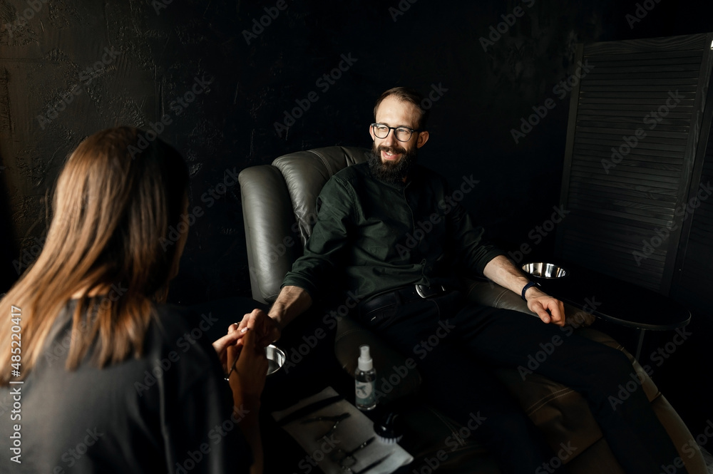 a satisfied man with a beard sits in a leather chair and is given a man's manicure. the concept of male manicure. a satisfied man with a beard sits in a leather chair and is given a man's manicure. the concept of male manicure.