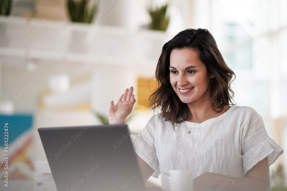 smiling remote working woman chatting and waving hand infront of a laptop or notebook during a video conference on her work desk in her modern bright living room home office smiling remote working woman chatting and waving hand infront of a laptop or notebook during a video conference on her work desk in her modern bright living room home office