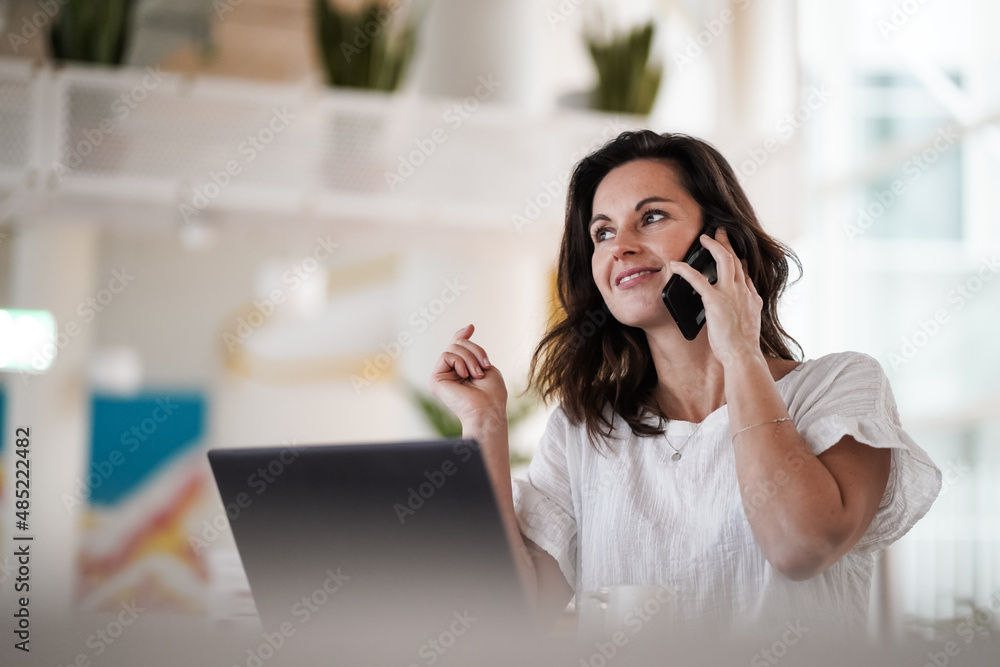 remote working dark haired woman calling and chatting infront of a laptop or notebook in casual outfit on her work desk in her modern airy bright living room home office with her mobile phone remote working dark haired woman calling and chatting infront of a laptop or notebook in casual outfit on her work desk in her modern airy bright living room home office with her mobile phone