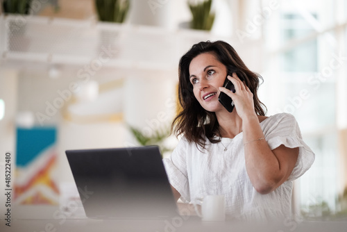 remote working dark haired woman calling and chatting infront of a laptop or notebook in casual outfit on her work desk in her modern airy bright living room home office with her mobile phone