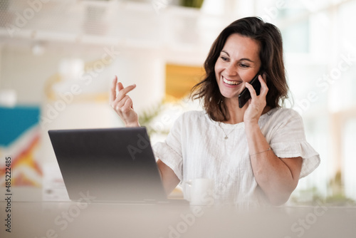 remote working dark haired woman calling and chatting infront of a laptop or notebook in casual outfit on her work desk in her modern airy bright living room home office with her mobile phone