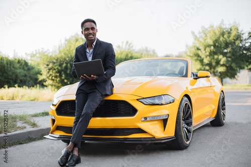Handsome African American man in a business suit. Businessman working with laptop. Man standing near his yellow sport car on the background of green park © sofiko14