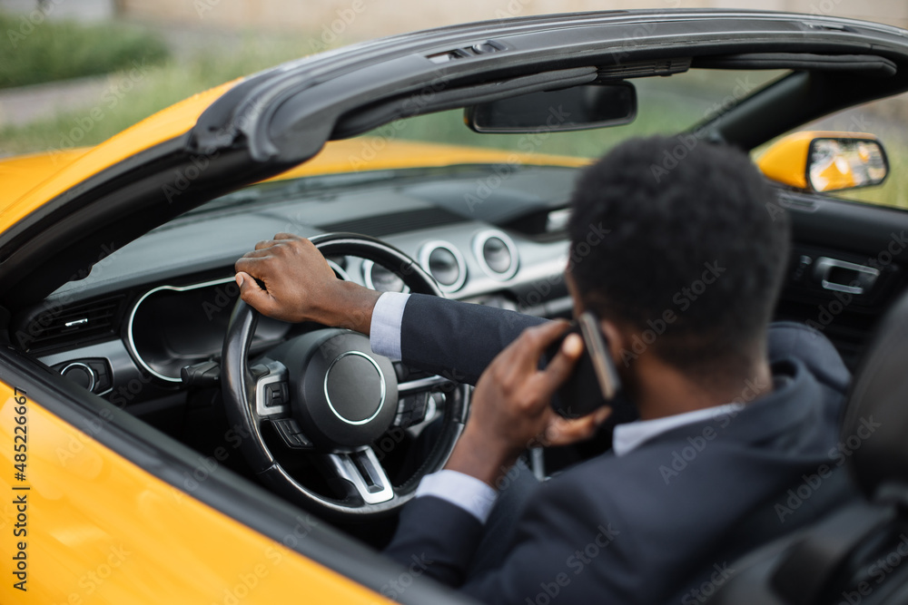 African Man in business suit using and talking on smartphone while driving a car, looking carefully at the road, back view.