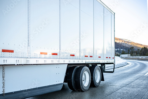 Big rig semi truck transporting cargo in dry van semi trailer with aerodynamic skirt driving on the winding winter slippery highway road with snow and ice