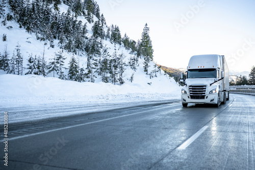 Bonnet big rig white semi truck transporting cargo in dry van semi trailer running on the winter dangerous slippery road with snow and ice in Montana