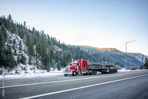 Powerful red classic big rig semi truck transporting industrial cargo on the long flat bed semi trailer climbing uphill on the wide winter highway road with snow and ice in Montana