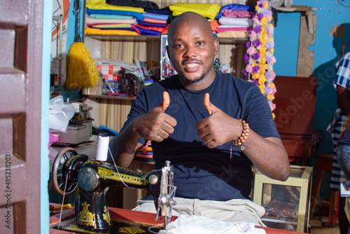 A happy African Nigerian male tailor, fashion designer or business man doing thumbs up gestures while making dress with a sewing machine in a tailoring shop