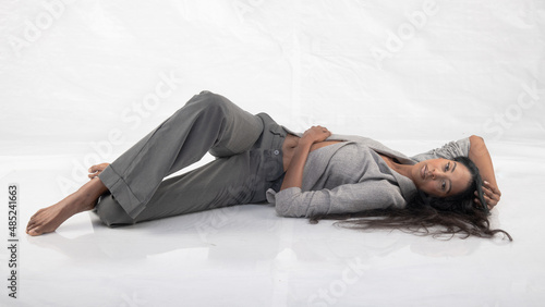 Tall glamorous woman in grey pants and jacket, reclining barefoot in a white studio setting.