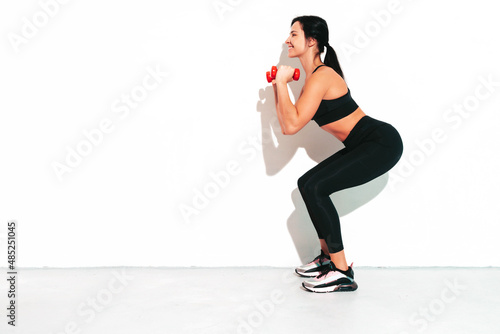 Fitness confident woman in black sports clothing. Sexy young beautiful model with perfect body. Female isolated on white wall in studio. Stretching out before training.Making squats with dumbbells