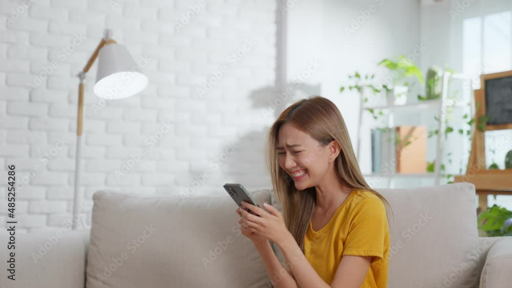 Smiling Asian woman sitting on the sofa looking at the screen on her mobile phone. Female received good news in an e-mail message. The excited woman stood up to celebrate and jumped in joy. Smiling Asian woman sitting on the sofa looking at the screen on her mobile phone. Female received good news in an e-mail message. The excited woman stood up to celebrate and jumped in joy.