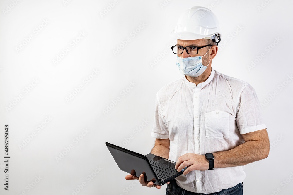 A male engineer with a netbook and a white hard helmet. Middle-aged man in a white shirt, glasses and a medical mask. Isolated portrait on a white background. Male stock photo. Copy space A male engineer with a netbook and a white hard helmet. Middle-aged man in a white shirt, glasses and a medical mask. Isolated portrait on a white background. Male stock photo. Copy space