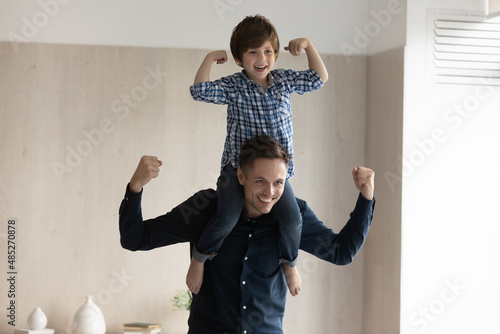Fototapeta Naklejka Na Ścianę i Meble -  Positive little son sitting on father shoulder, showing strength gesture, smiling loving young dad and 6s boy child engaged in funny activity at home, playing superheroes, happy fathers day concept