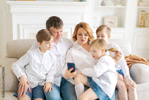Happy parents with three kids using smartphone at home together sitting on cozy sofa in living room, smiling mother and father with two sons and daughter looking at phone screen taking selfie cheerful