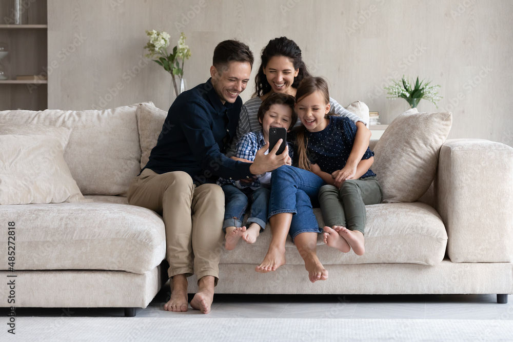 Happy parents and children having fun with smartphone at home, smiling mother and father with kids looking at smartphone screen, sitting on cozy couch, watching cartoons or making video call Happy parents and children having fun with smartphone at home, smiling mother and father with kids looking at smartphone screen, sitting on cozy couch, watching cartoons or making video call