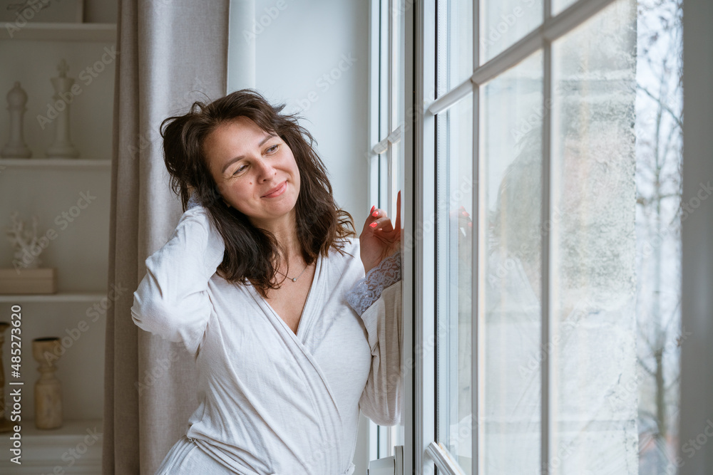 Happy and cheerful woman of Caucasian ethnicity in a light robe stands by the window in the morning and smiles