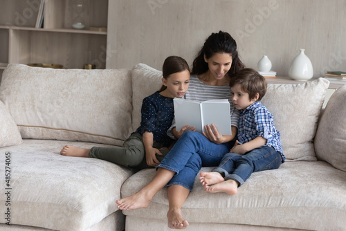 Loving mother reading book to little son and daughter, sitting relaxing on cozy couch at home, caring young mom telling to kids funny fairy tale story, spending leisure time weekend together © fizkes