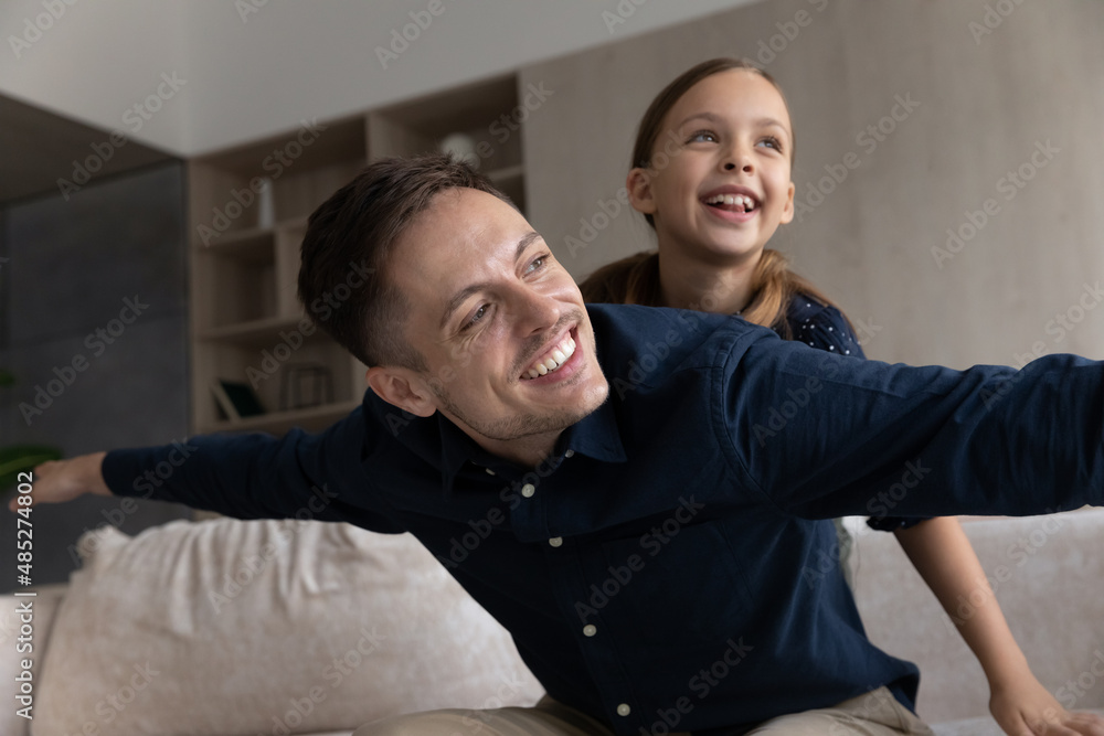 Obraz premium Smiling father piggy backing daughter, looking in distance, dreaming and having fun at home, happy loving young dad pretending plane with hands outstretched, family engaged in funny activity
