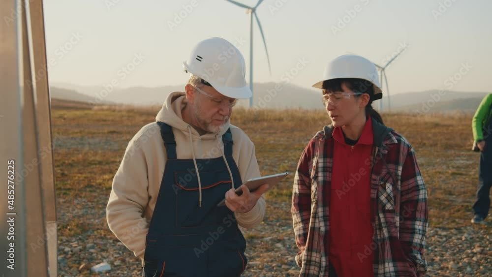 Portrait engineers in hardhats stand using tablet computer to check wind turbine system working at sunset on wind power station. Slow motion Portrait engineers in hardhats stand using tablet computer to check wind turbine system working at sunset on wind power station. Slow motion