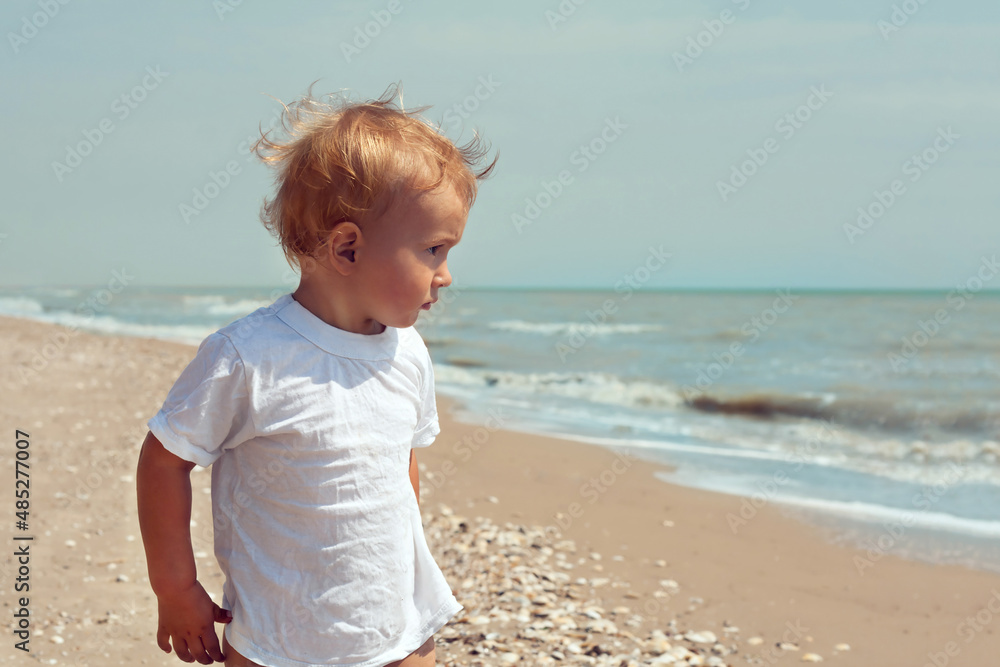 A little boy of 2 years in a white T-shirt and fluffy hair looks seriously at the sea with water and sand on the background. Free place for copy space A little boy of 2 years in a white T-shirt and fluffy hair looks seriously at the sea with water and sand on the background. Free place for copy space
