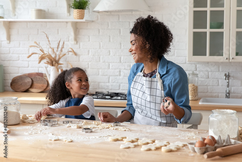 Joyful laughing small cute multiracial child daughter enjoying learning preparing homemade cookies with African American mother or babysitter, using cutters on dough, involved baking in kitchen.