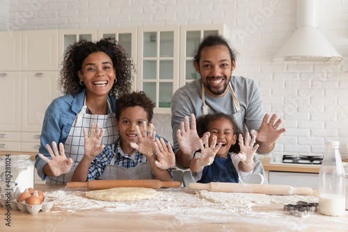 Portrait of happy African American young couple parents having fun with joyful small cute laughing children son daughter, enjoying cooking homemade pastry, pancakes or cookies in modern kitchen.