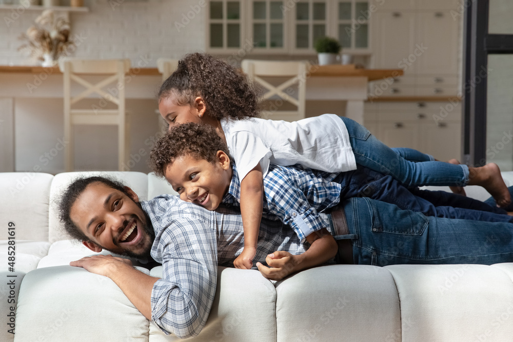 Joyful adorable laughing African American children son daughter lying on back of sincere smiling millennial father, playing together on big cozy sofa, enjoying family weekend playtime activity. Joyful adorable laughing African American children son daughter lying on back of sincere smiling millennial father, playing together on big cozy sofa, enjoying family weekend playtime activity.