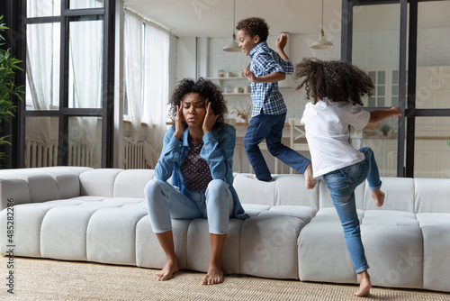 Unhappy depressed young African American mother or babysitter suffering from strong headache, annoyed by overjoyed hyperactive loud screaming kids son daughter playing running around in living room. photo
