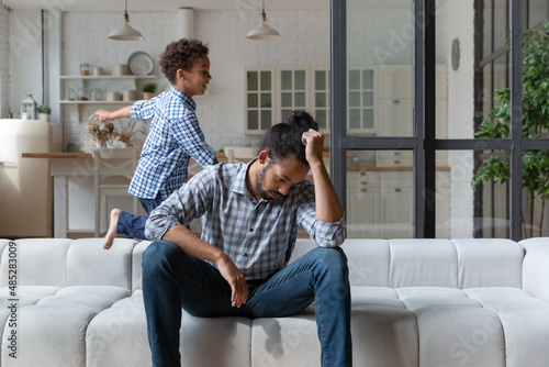 Vászonkép Depressed frustrated young African American father feeling tired, suffering from headache, irritated by noisy little child son playing having fun in living room, raising disobedient kids problems