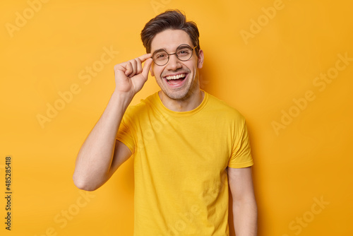 Waist up shot of cheerful brunet guy keeps hand on rim of spectacles wears casual t shirt and round spectacles being in good mood poses against vivid yellow background. Happy emotions concept