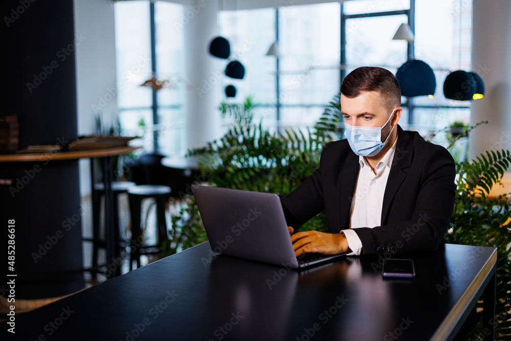 Handsome young man in a mask working in a cafe with a laptop during the coronavirus pandemic.