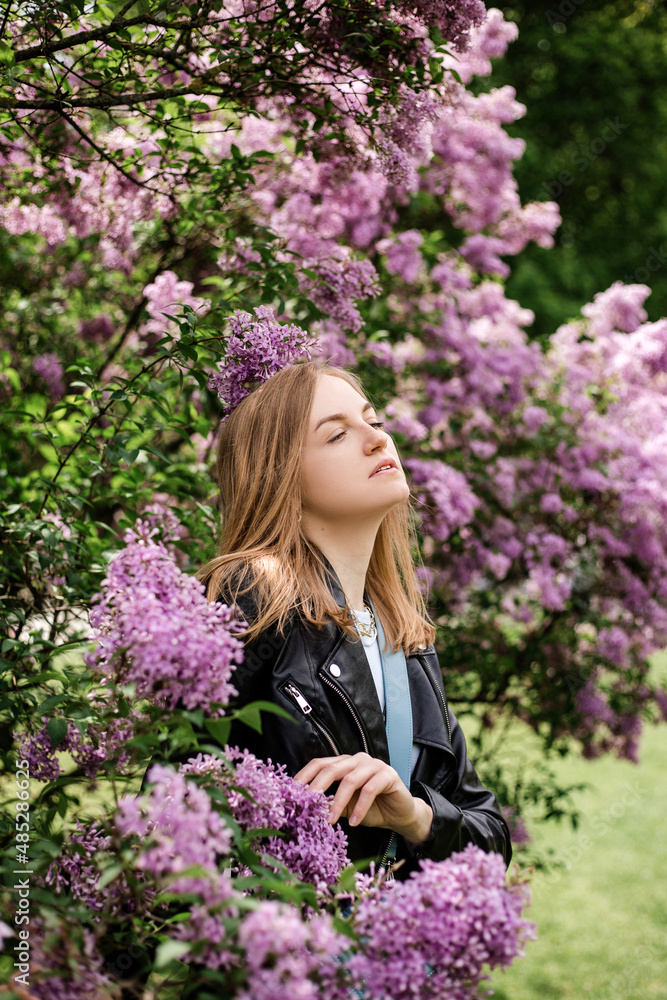 Young woman with blond hair wearing blue top and black jacket walking in spring park. Beautiful woman enjoying the smell of Lilac. Positive emotions and feelings. Holiday concept, Womans Day banner Young woman with blond hair wearing blue top and black jacket walking in spring park. Beautiful woman enjoying the smell of Lilac. Positive emotions and feelings. Holiday concept, Womans Day banner