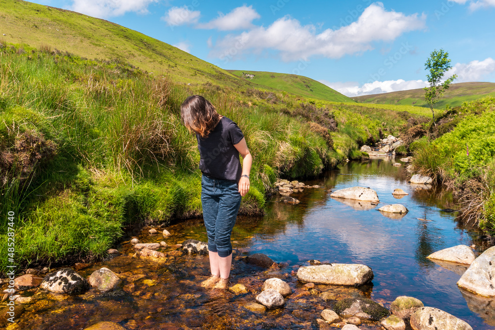 Naklejka premium Woman hiker standing barefoot in a cold mountain stream, in Wicklow Mountains, Ireland. Cooling off after hiking on a hot summer day.