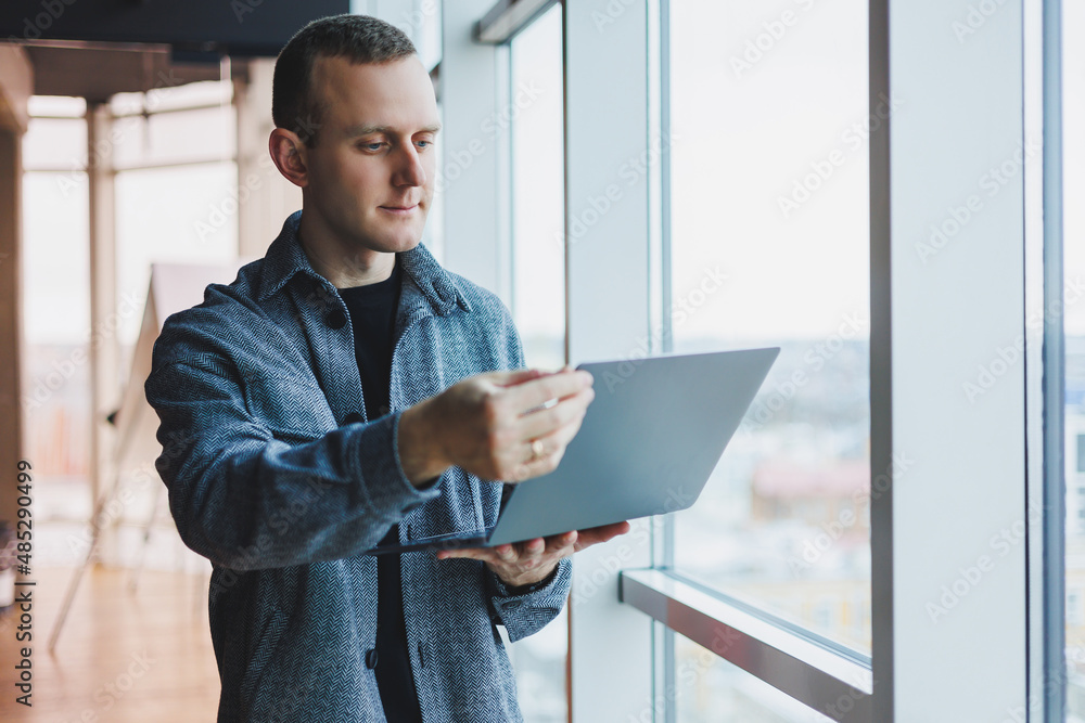 Portrait of a smart intelligent male manager, he is holding a laptop for research in the office, a business man in elegant clothes playing on the touchpad. Standing in the office by a large window Portrait of a smart intelligent male manager, he is holding a laptop for research in the office, a business man in elegant clothes playing on the touchpad. Standing in the office by a large window