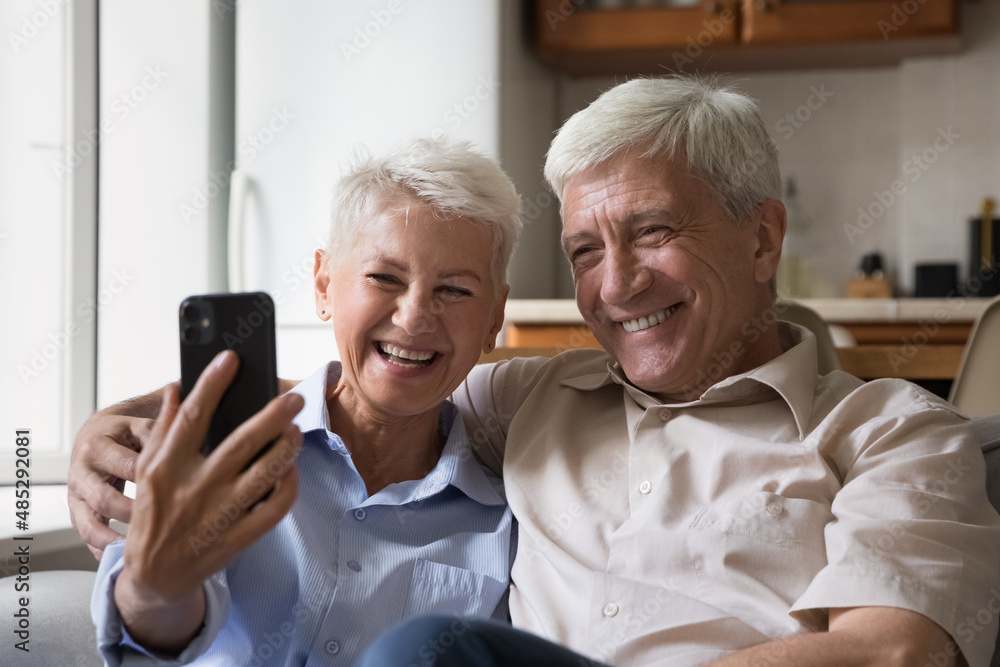 Happy joyful senior couple taking home selfie on mobile phone, posing for photo with toothy smile, resting on couch with digital gadget, hugging, having fun, laughing, making video call on smartphone Happy joyful senior couple taking home selfie on mobile phone, posing for photo with toothy smile, resting on couch with digital gadget, hugging, having fun, laughing, making video call on smartphone