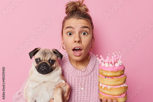 Shocked woman poses with cut dog celebrates first birthday of pet holds doughnutss surprised to hear unexpected news poses against pink background. Domestic animals people and celebration concept