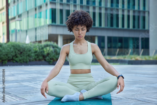 Relaxed fit woman in tracksuit sits in lotus pose on karemat breathes deeply practices yoga tries meditates outdoors poses against modern city building. Healthy living and relaxation concept