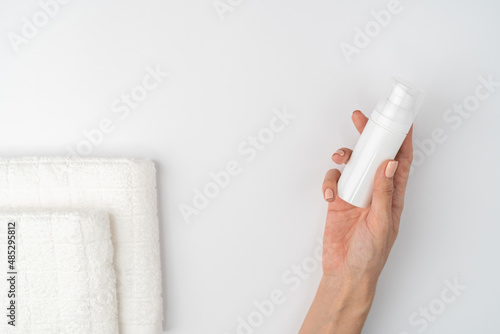A close shot of the thin graceful hands of a girl with a nude manicure holds a bottle of foam for washing, cleansing the skin of the face, on a white background. Fresh towel.