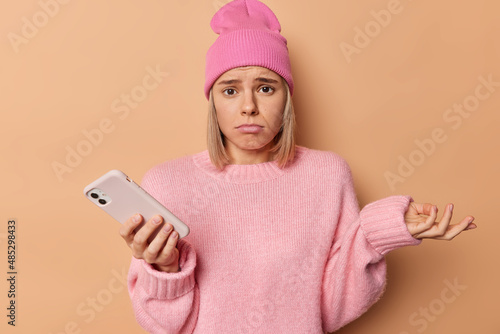 Hesitant confused displeased woman shrugs shoulders looks frustrated at camera doesnt know how to make payments online scrolls social media wears pink hat and jumper isolated over brownn background. photo