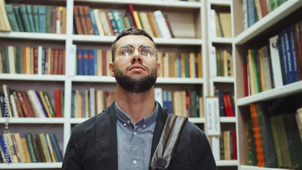 Bearded young man with eyeglasses wearing shirt and jacket standing among bookcases and looking up. Student choosing book in library. Concept of education Bearded young man with eyeglasses wearing shirt and jacket standing among bookcases and looking up. Student choosing book in library. Concept of education