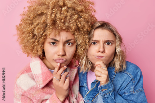 Serious young women look attentively at camera hold chins purse lips concentrated forward wear fashionable clothes stand closely to each other against pink background listen something attentively