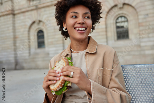 Принти на полотні Happy curly haired woman sits outdoors eats delicious sandwich looks gladfully away prefers junk food dressed in casual clothes has lunch break after strolling in city