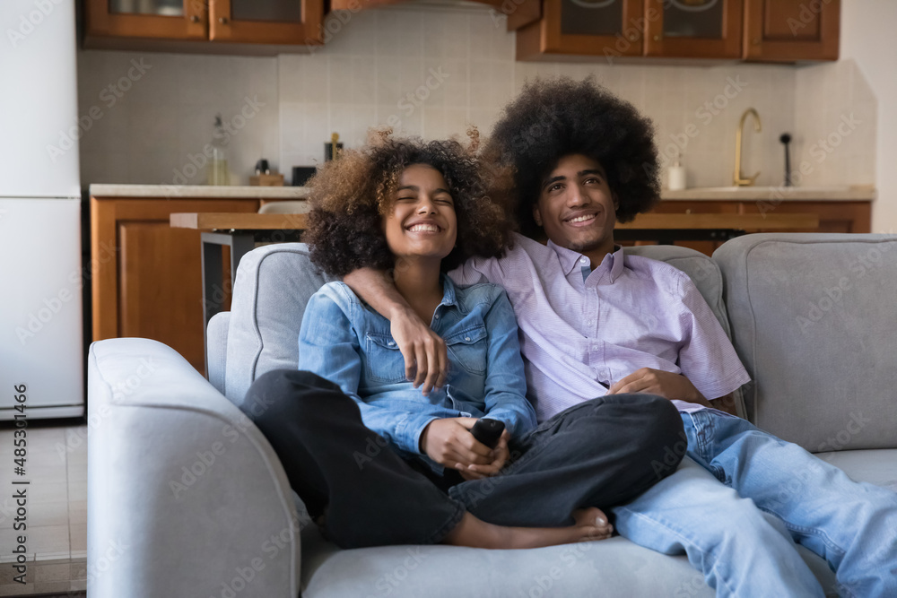 Happy joyful teen African couple watching TV, comedy movie, sitcom series, resting on sofa in living room, using remote control, hugging, laughing, enjoying home leisure, multimedia entertainment Happy joyful teen African couple watching TV, comedy movie, sitcom series, resting on sofa in living room, using remote control, hugging, laughing, enjoying home leisure, multimedia entertainment