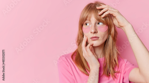 Horizontal shot of thoughtful redhead teenage girl keeps hand on face and head applies hydrogel patches under eyes undergoes beauty procedures has freckled skin isolated over pink background